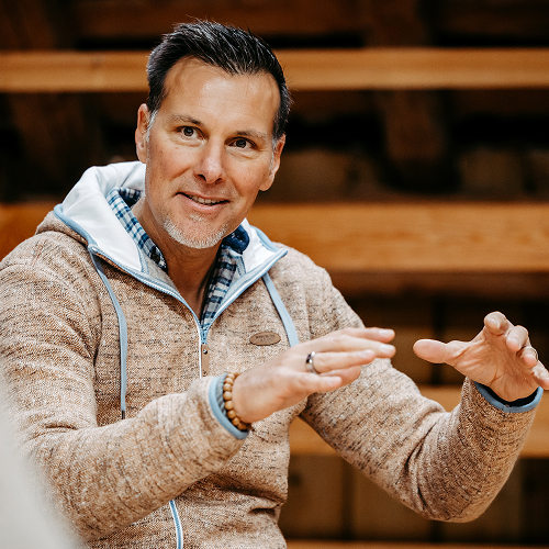 Ein Mann mit kurzen dunklen Haaren und einem gestutzten Bart gestikuliert mit seinen Händen, während er spricht. Er trägt eine beigefarbene Jacke über einem blau karierten Hemd und sitzt vor einem hölzernen Hintergrund.