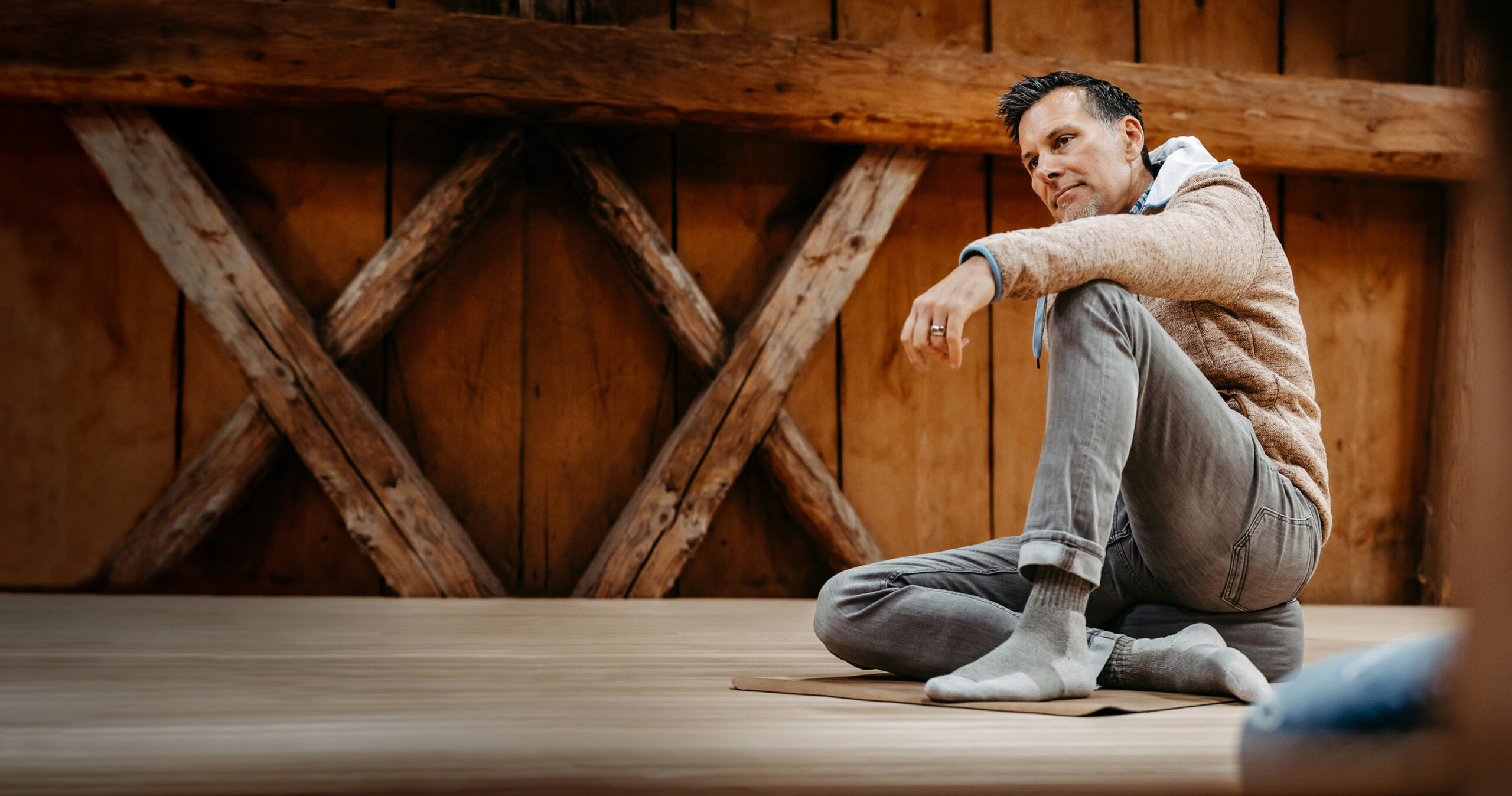 Ein Mann mit Kapuzenpulli und Socken sitzt auf einer Yogamatte in einem Innenraum und dreht seinen Körper in einer sitzenden Yogapose, während im Hintergrund eine Holzwand und gekreuzte Balken zu sehen sind.