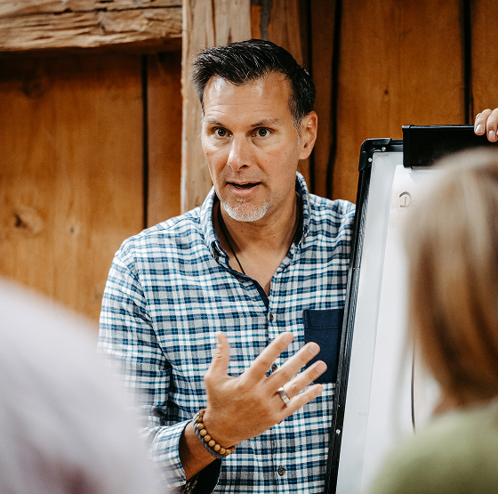 Ein Mann in einem karierten Hemd gestikuliert vor einem Flipchart und spricht zu einer kleinen Gruppe von Menschen in einem rustikalen, hölzernen Raum.