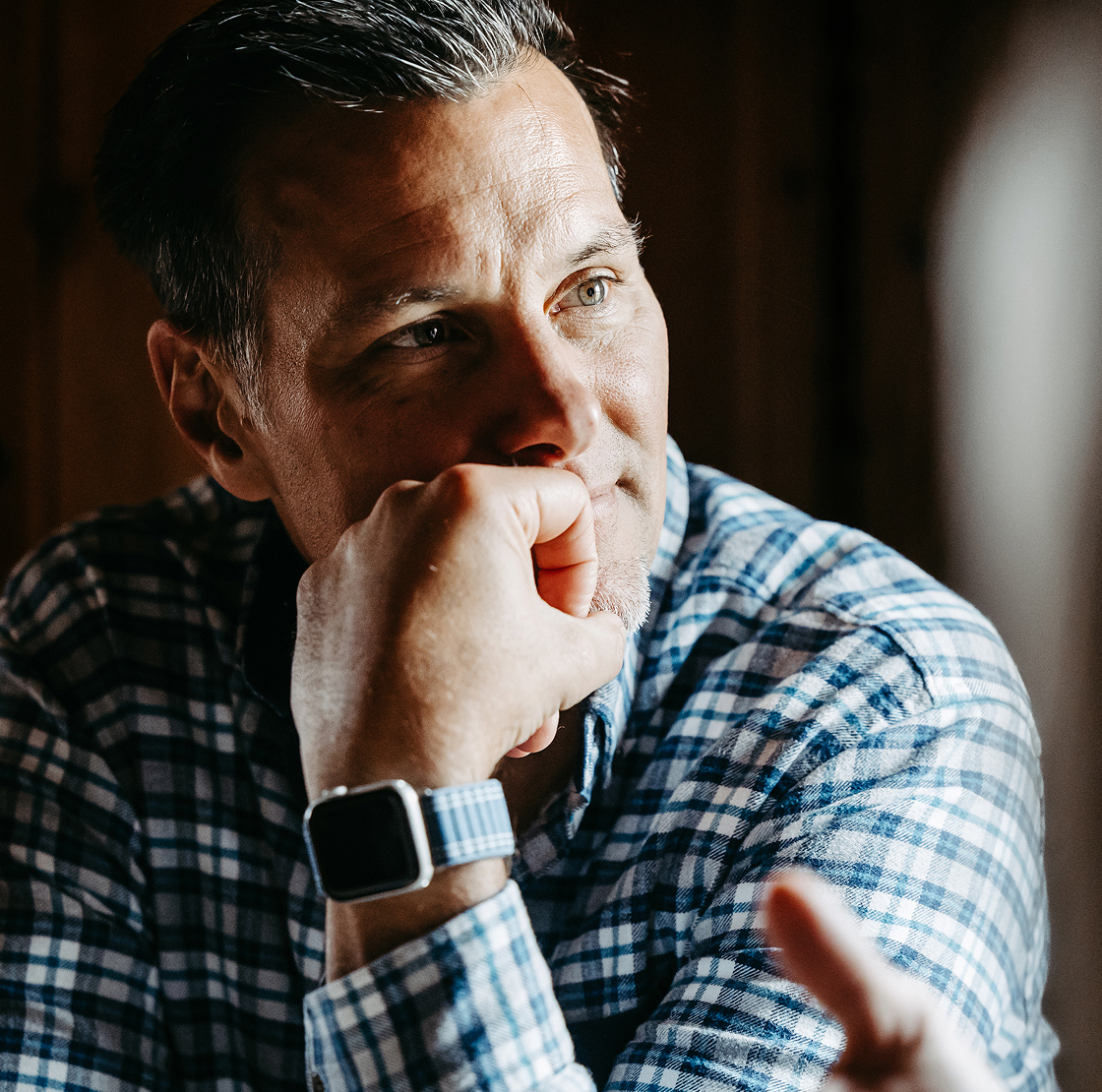 Ein Mann in einem blau karierten Hemd stützt sein Kinn auf seine Hand und wirkt nachdenklich. Er trägt eine Smartwatch und blickt in Richtung einer Person, deren Hand im Vordergrund zu sehen ist, was auf ein Gespräch hindeutet.